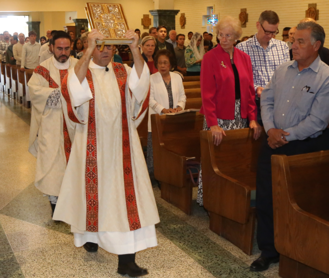 Priest in white robe carries a book through a church aisle during a procession