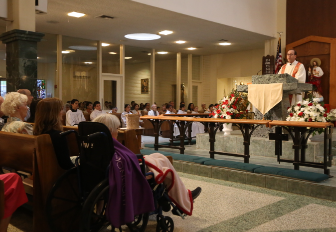 People seated in a church for a memorial service, with a speaker at the podium and flowers on the altar.