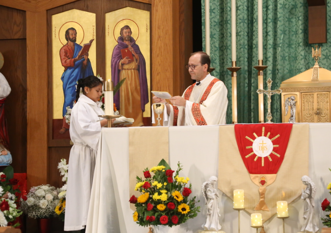 Priest and altar server at church altar during Mass, with candles, flowers, and religious icons behind them