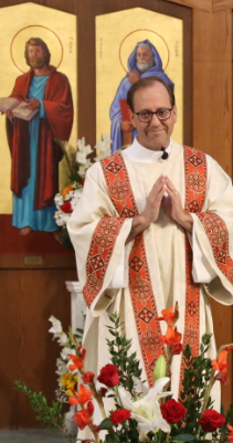 Priest in white and red vestments stands in church altar with hands joined in prayer, flowers and saints behind him