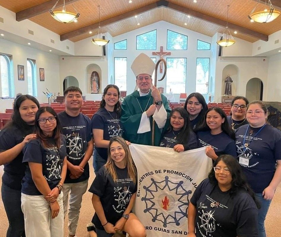 Group of people posing in a church, smiling with a bishop and holding a banner beneath a crucifix.