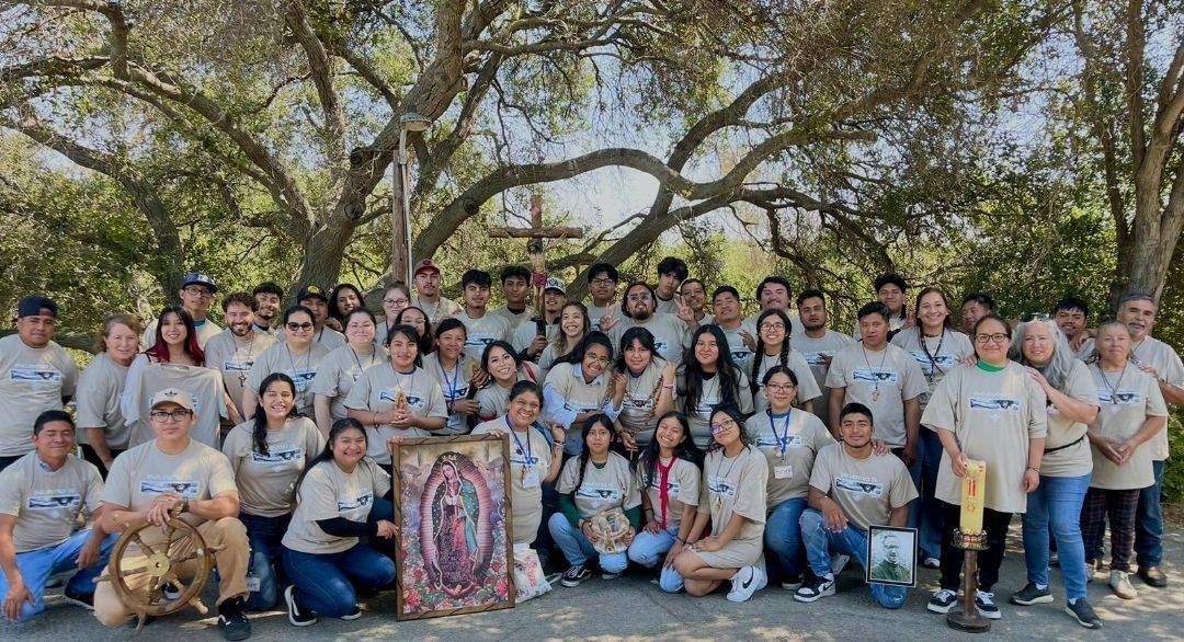 Large group posing outdoors under trees, with a colorful portrait banner at the center.