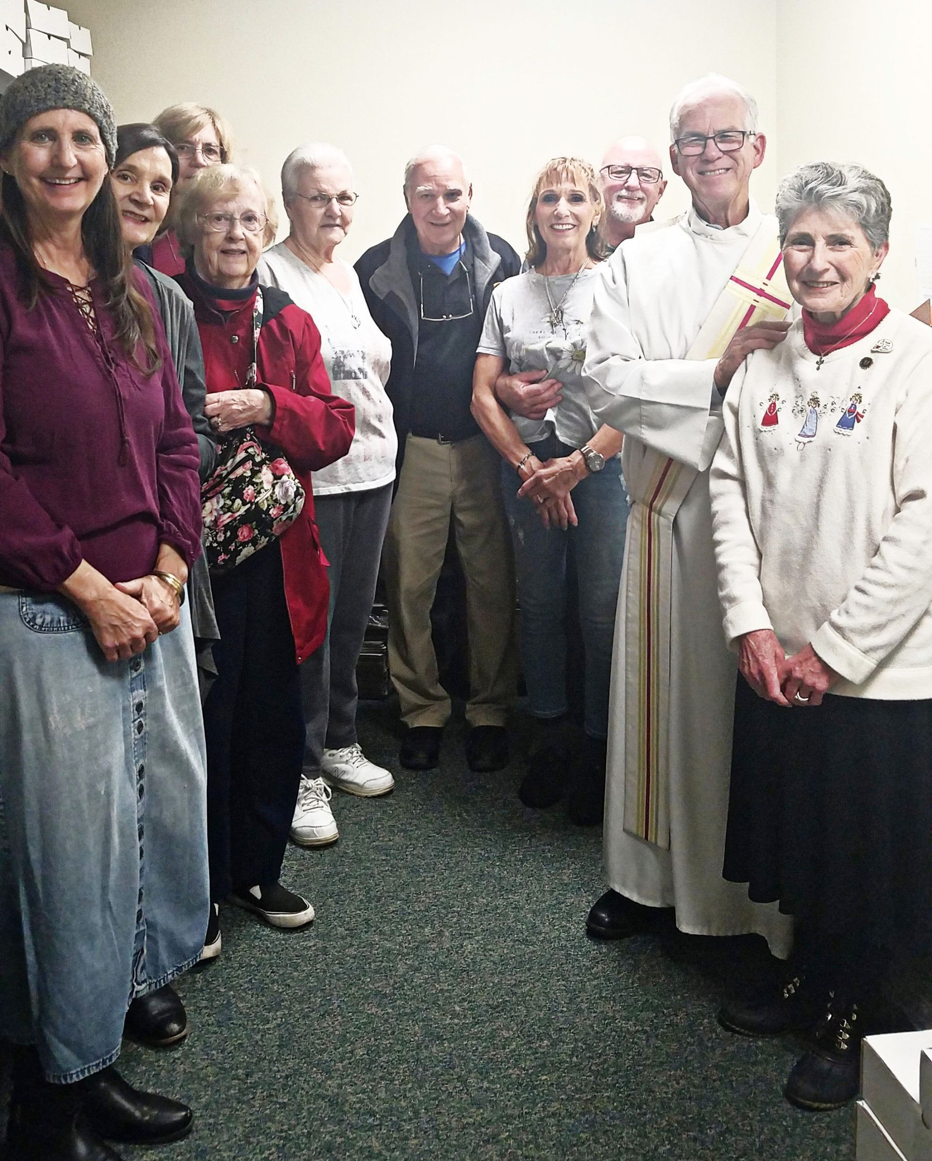 Group of people standing together indoors, some holding candles, in a hallway with white walls and carpet.