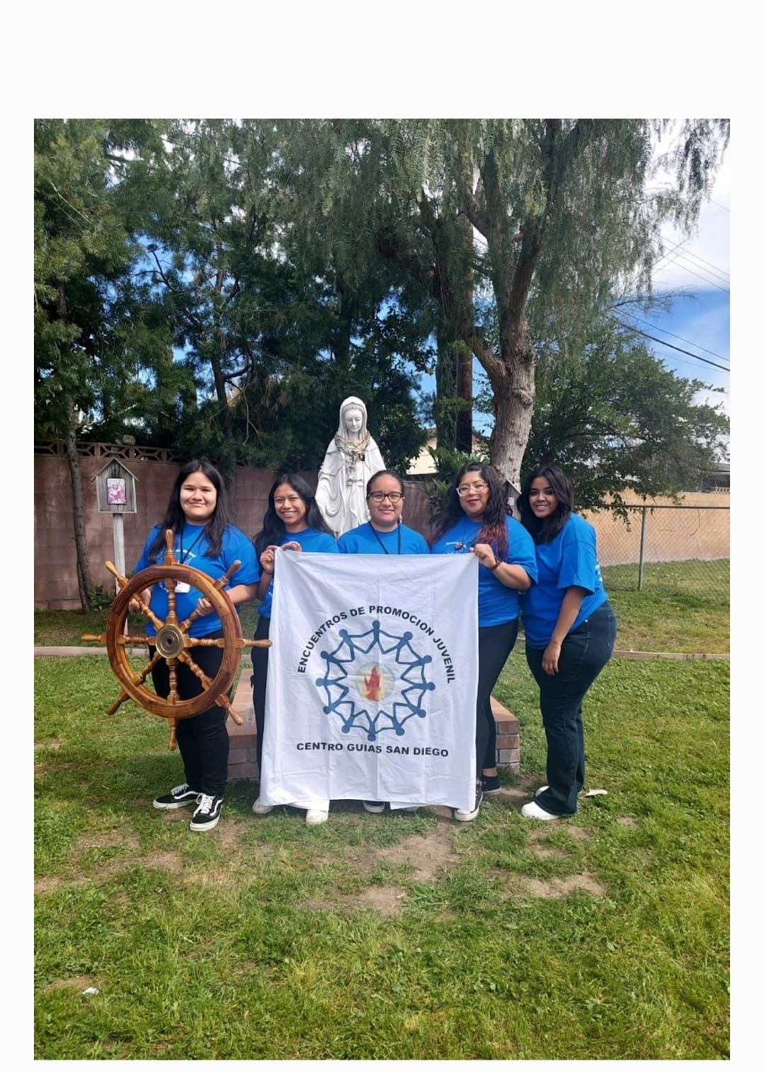 Five people in blue shirts posing outdoors with a ship wheel and banner in front of a tree.