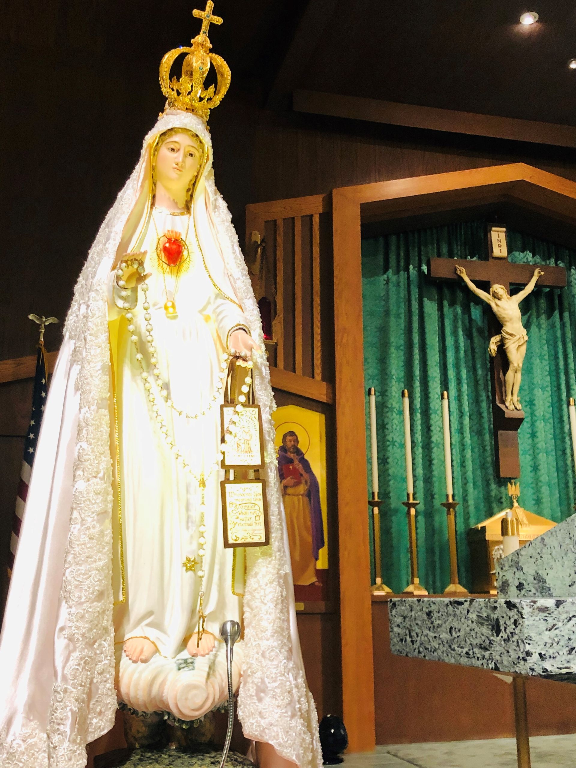 Statue of the Virgin Mary in a white robe inside a church, with a crucifix on the wall nearby.