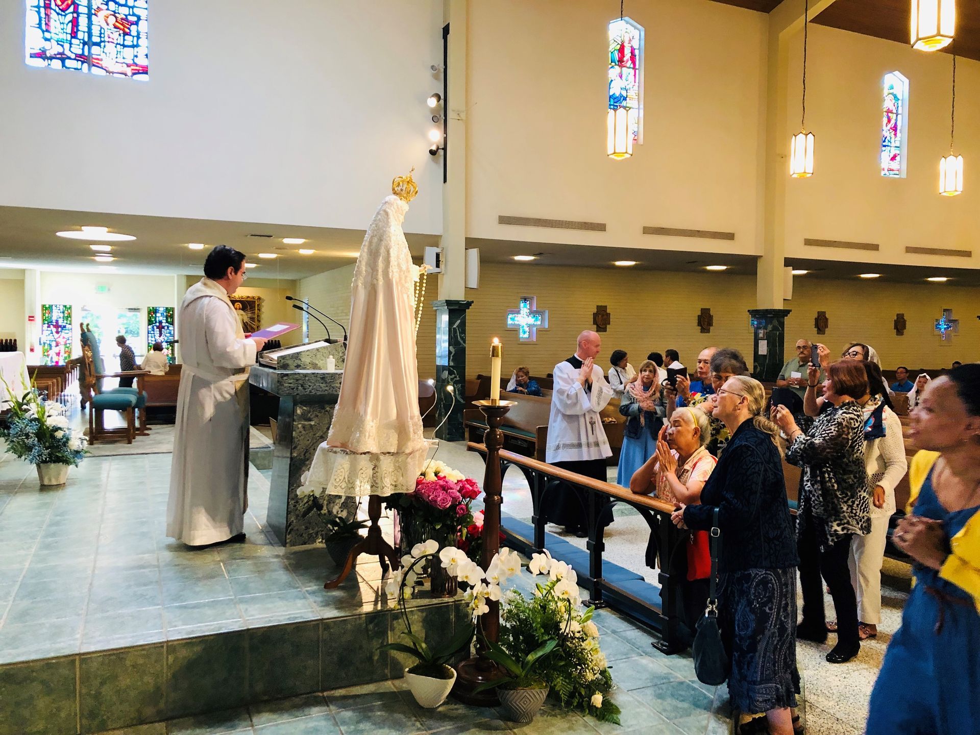 Church ceremony with priest at the altar and congregation standing beside a statue of Mary and flowers.