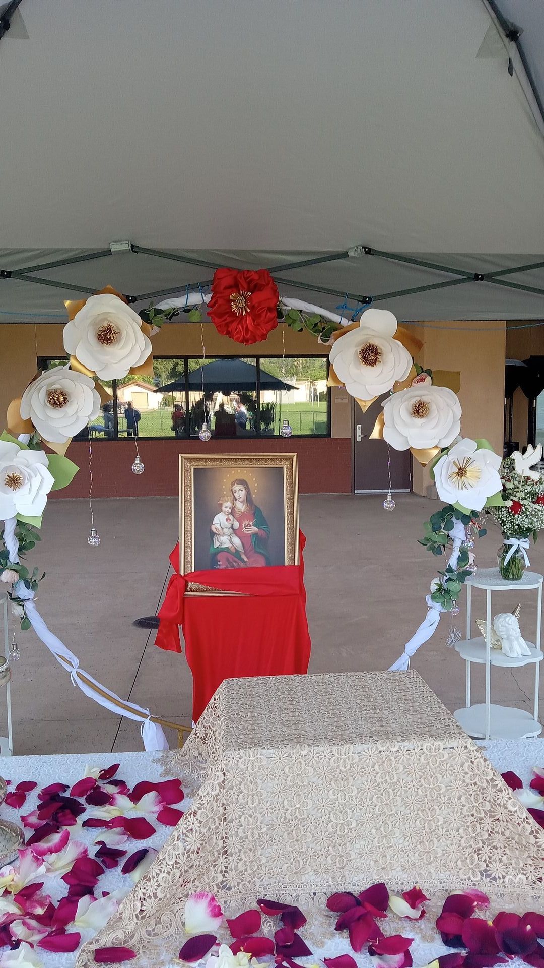 Memorial altar with framed portrait, white floral arch, red table, and rose petals in a covered outdoor space