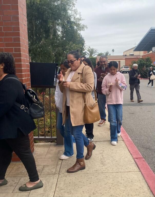 People standing in a line outside a brick building, some checking phones and wearing coats.