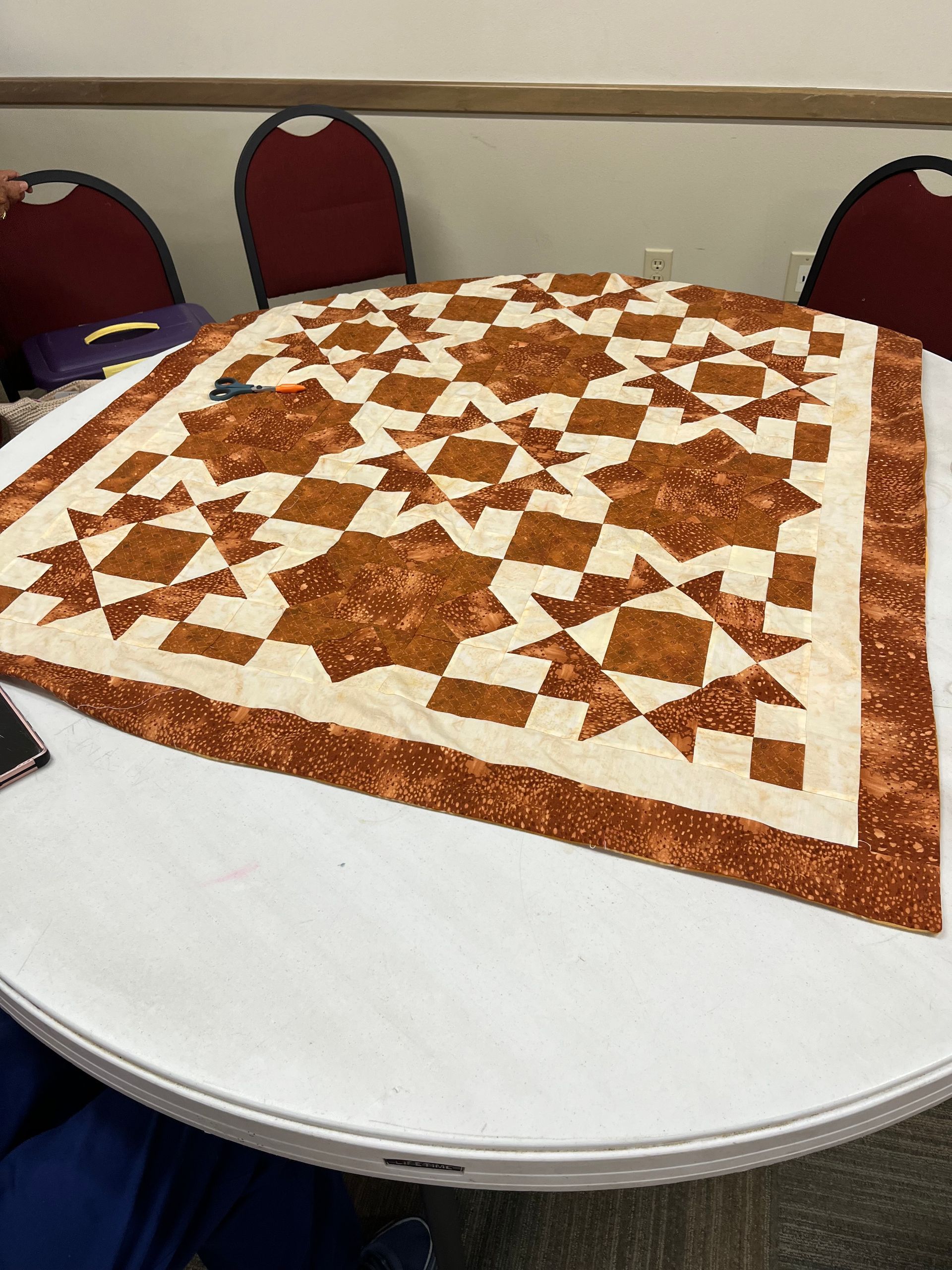Brown and white quilt spread across a round table with red chairs in the background