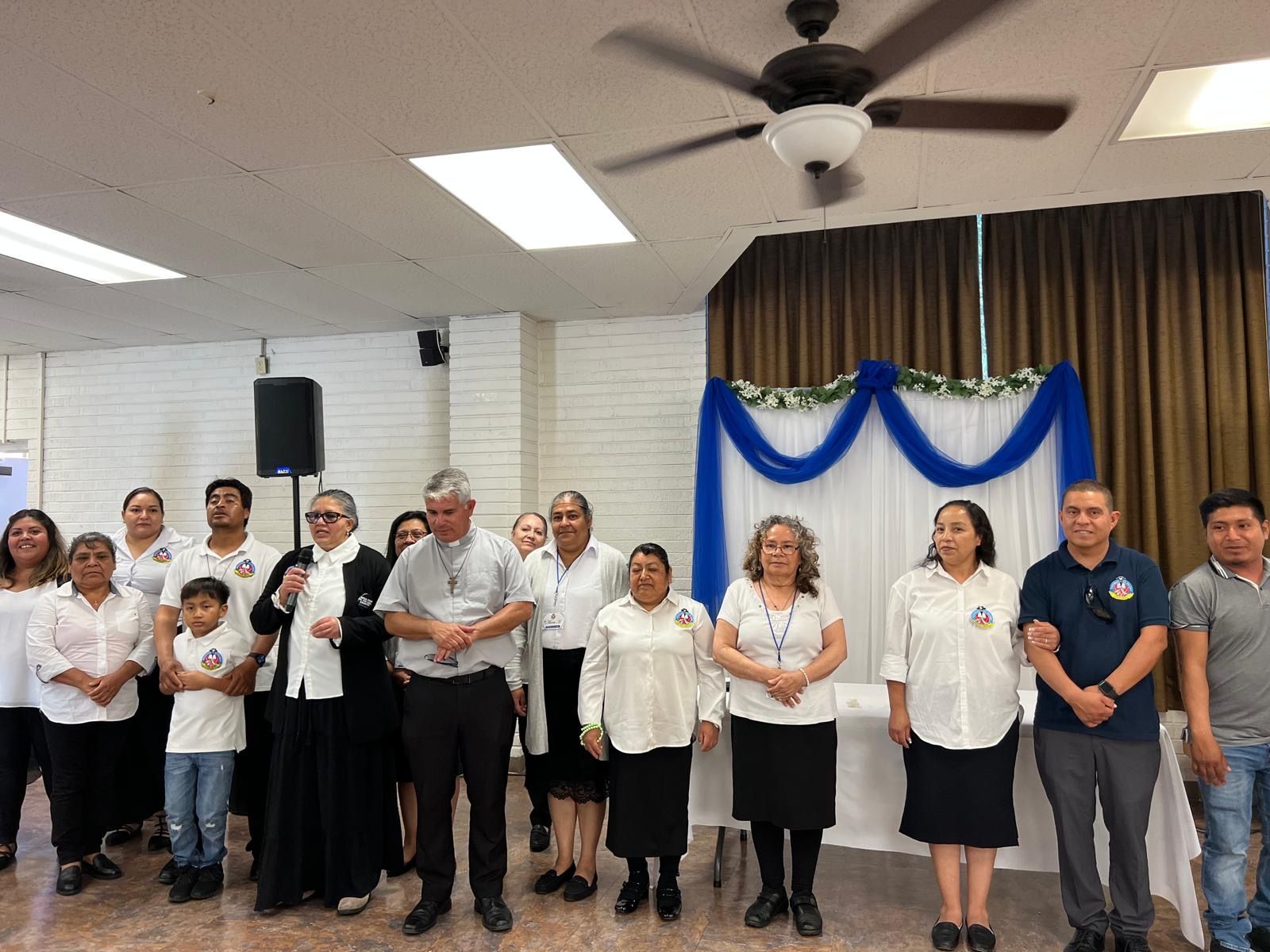 Group of people posing indoors at a ceremony, with blue drapes on the wall and a ceiling fan overhead.
