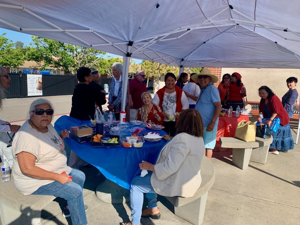 People seated around a blue-covered table under a white canopy at an outdoor gathering.