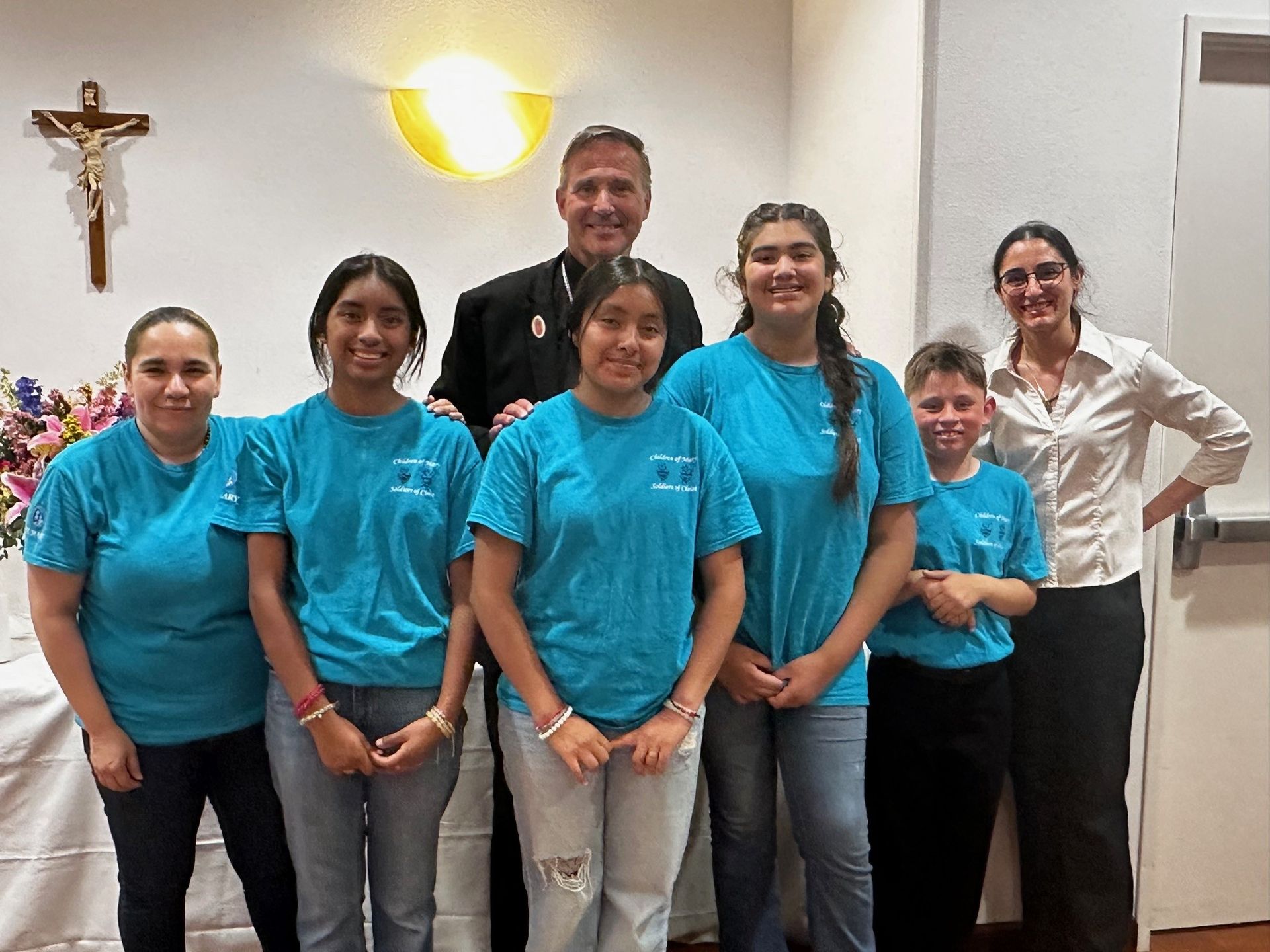 Group of people posing indoors, wearing turquoise shirts, with a crucifix and wall light behind them.