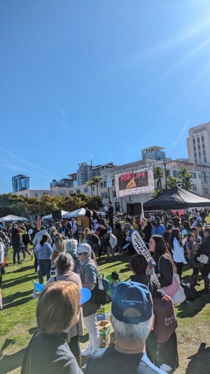 Crowd gathered in a sunny park at an outdoor festival with tents and a stage in the background.