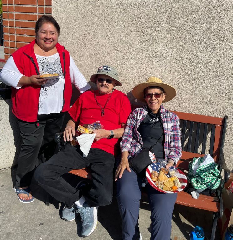 Three people in red tops sit and stand by a bench outdoors, smiling and holding food containers.
