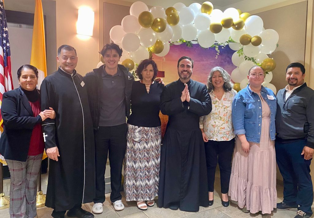 Eight people standing together indoors under a white, gold, and black balloon arch, smiling at a celebration.