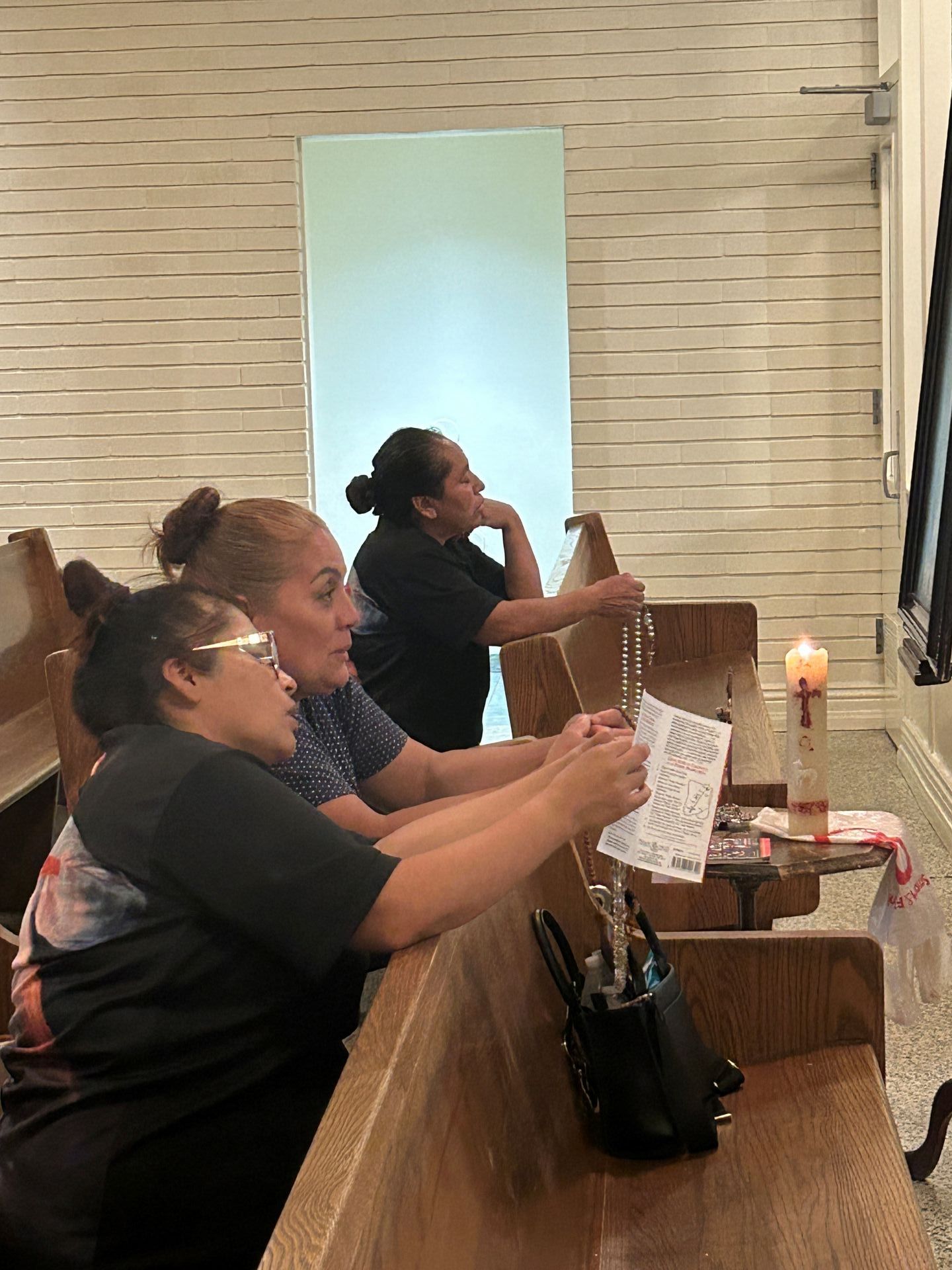 People seated in church pews, one holding a paper, with a lit candle on a small table nearby.