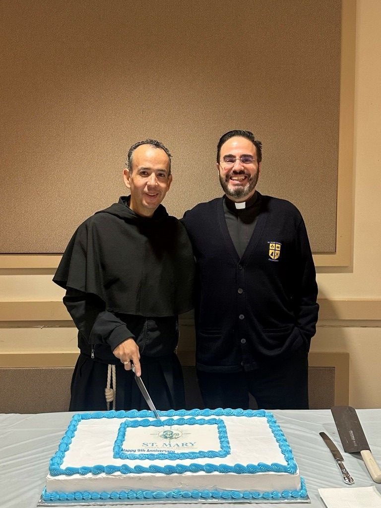Two men in black stand behind a blue-and-white cake, smiling at a table.