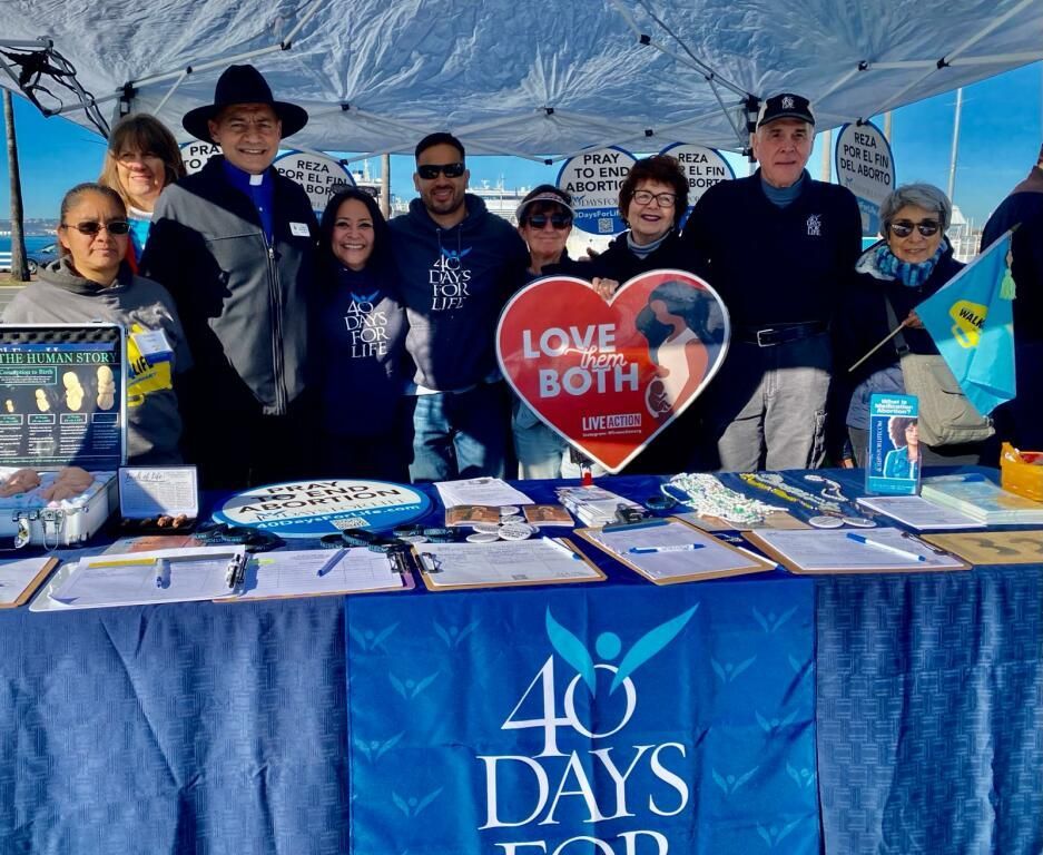 Group behind a blue table at a 40 Days for Life booth, holding a heart sign reading “Love Both.”