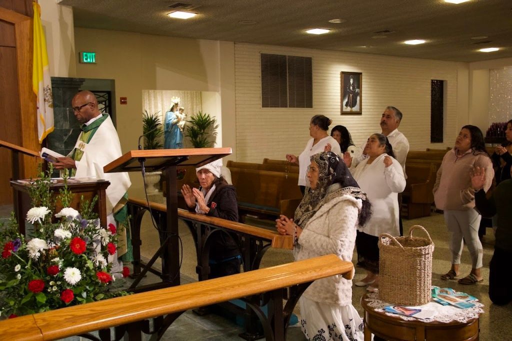 People standing in a church during a service, with a priest at the altar and flowers in front.