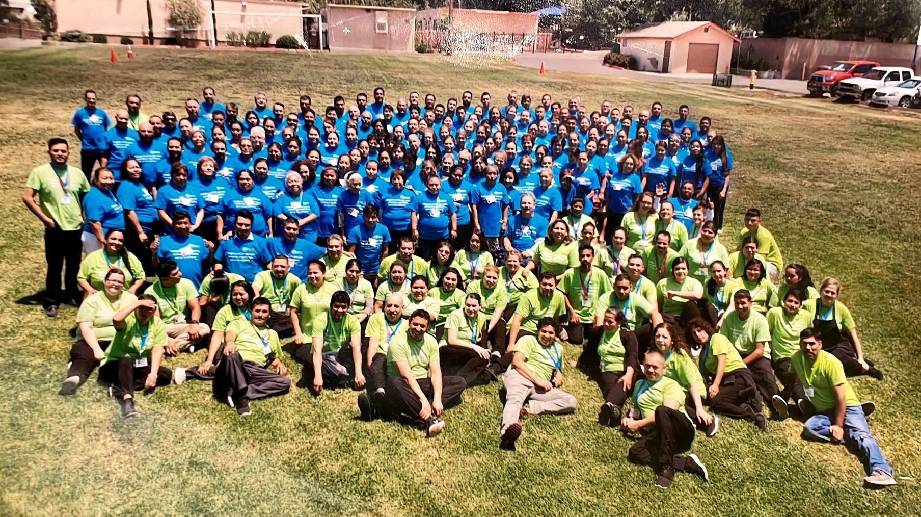 Large group in blue and green shirts posing for a team photo outdoors on a grassy field.