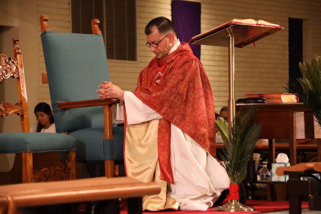 Priest in red vestments kneels beside a blue chair during a church service