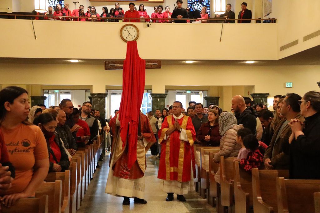 Ceremonial procession in a hall with red robes and a tall red banner, flanked by seated audience on both sides