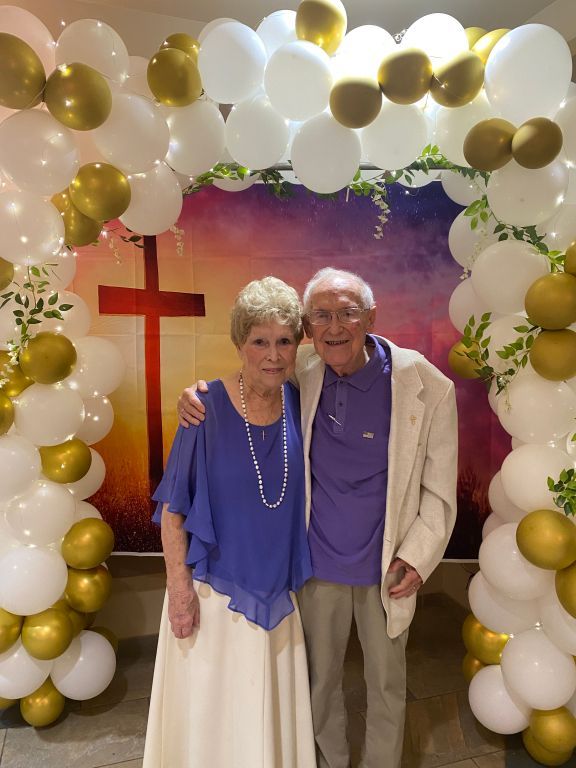 Two people smiling in front of a white, gold, and green balloon arch with a red cross backdrop