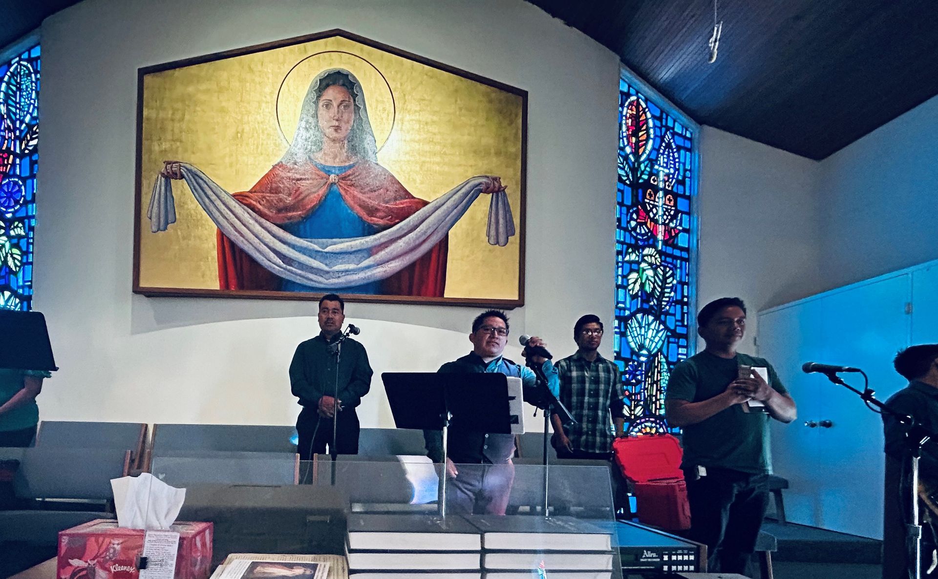 Band performing on a church stage beneath a large Jesus mural, with microphones and instruments.