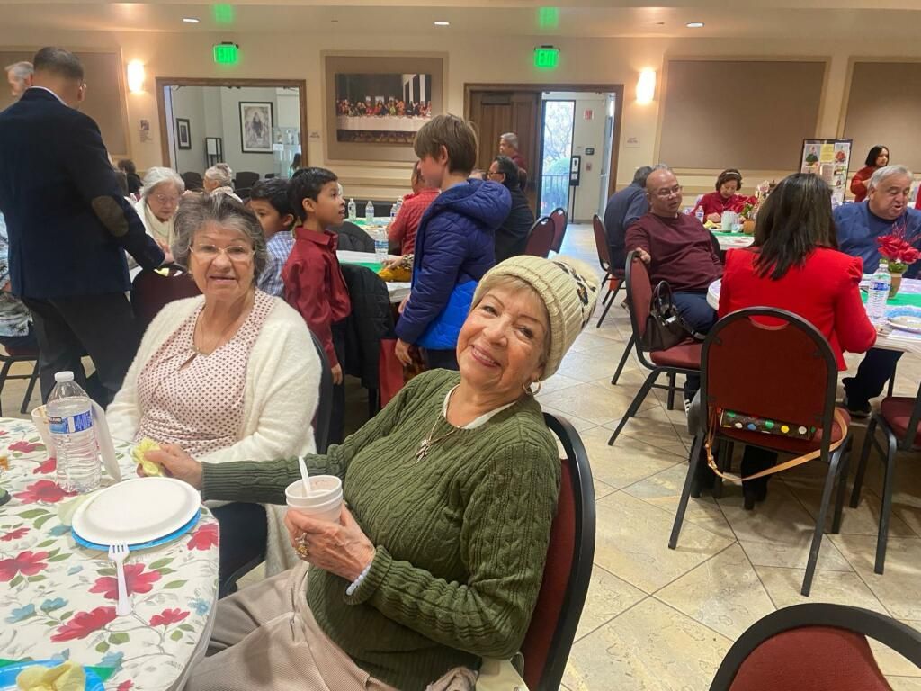 People seated at tables in a bright banquet hall, smiling and chatting during a community gathering