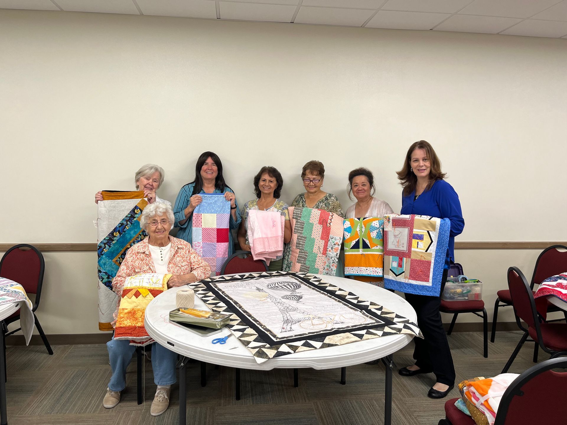 Group posing behind a decorated table with quilted bags and a large black-and-white cake in a meeting room