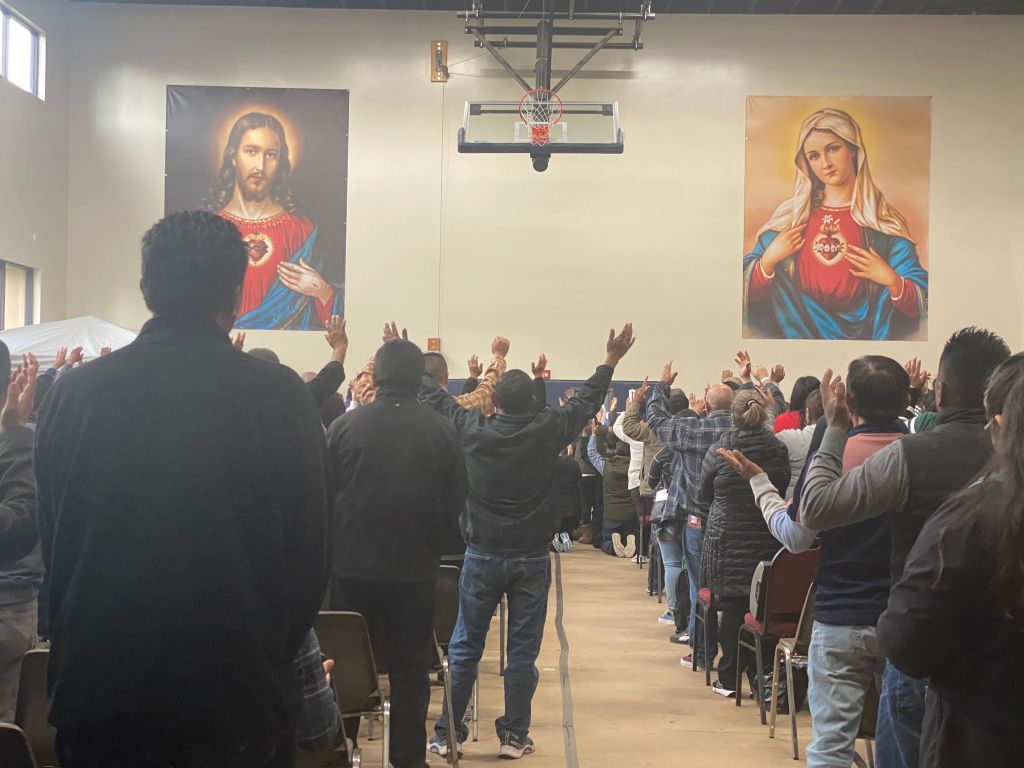 People in a church with raised hands during worship, with Jesus and Mary portraits on the walls.