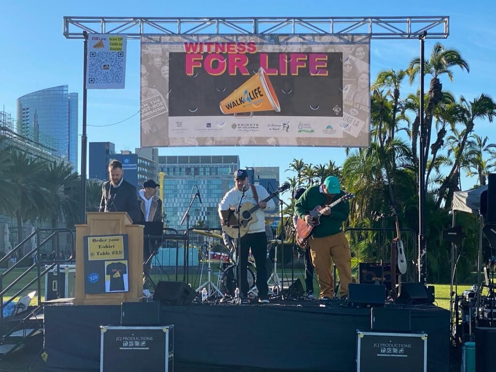 Outdoor live band performing on a stage under a “For Life” banner, with palm trees and city buildings behind.