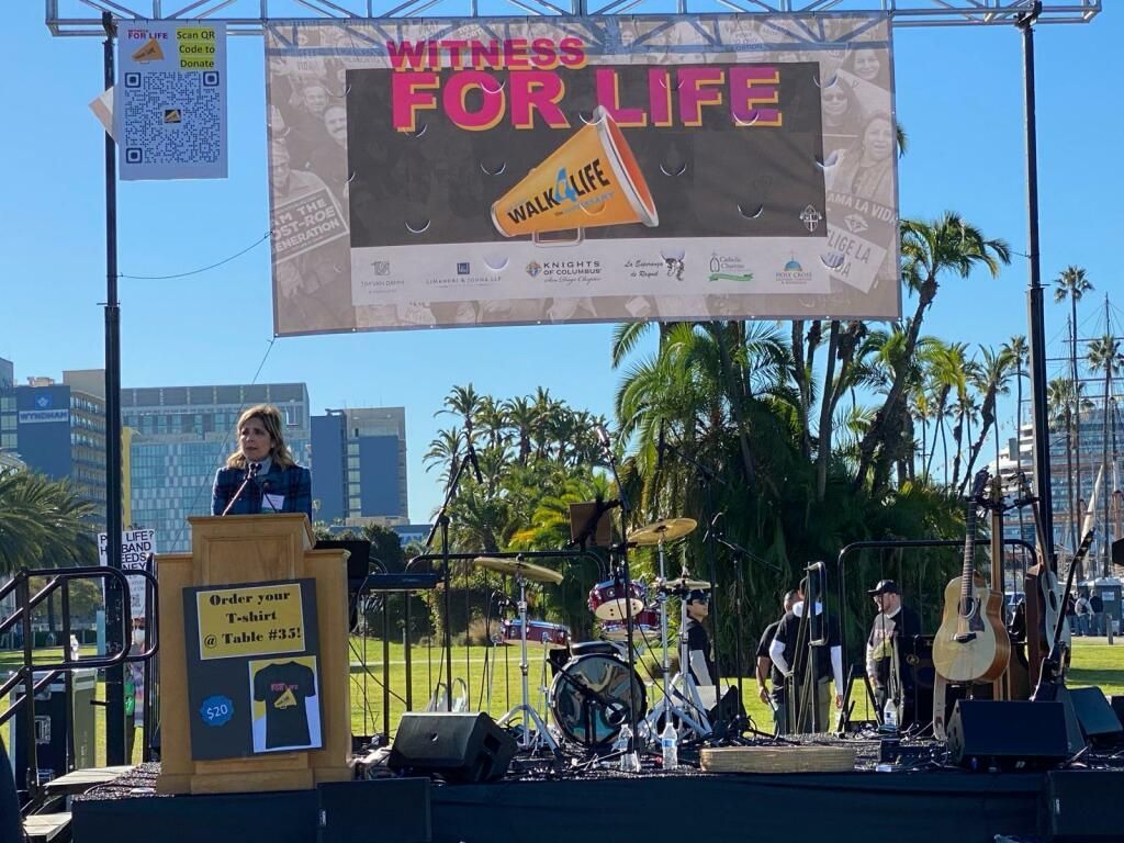 Outdoor rally stage with a speaker at a podium under a “Wins for Life” banner, guitars and crowd visible.