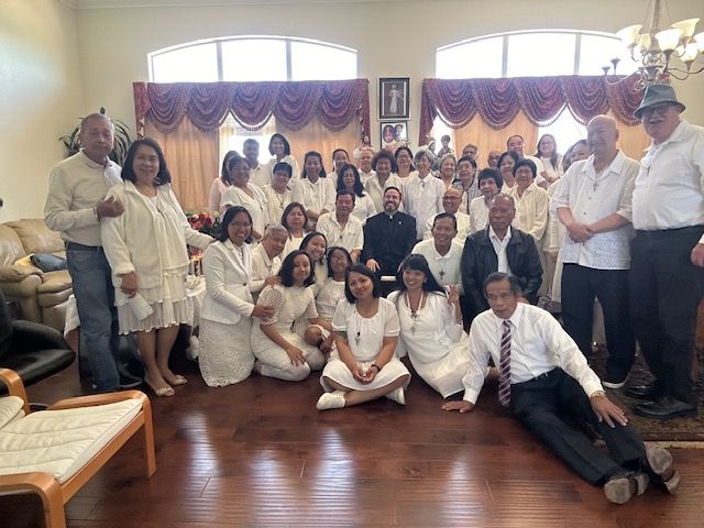 Large group posing in a living room with wooden floors, cream curtains, and framed wall art.