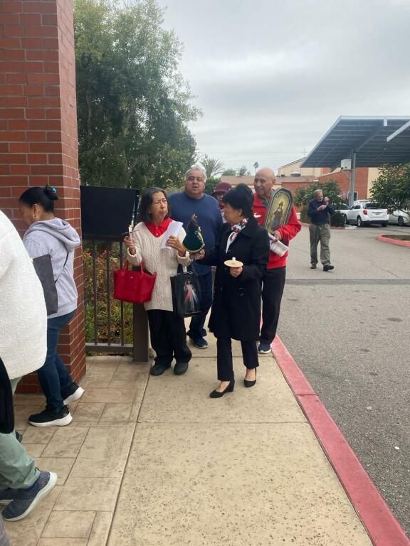 People gathered outside a building, standing on a sidewalk near a brick wall and parking lot.