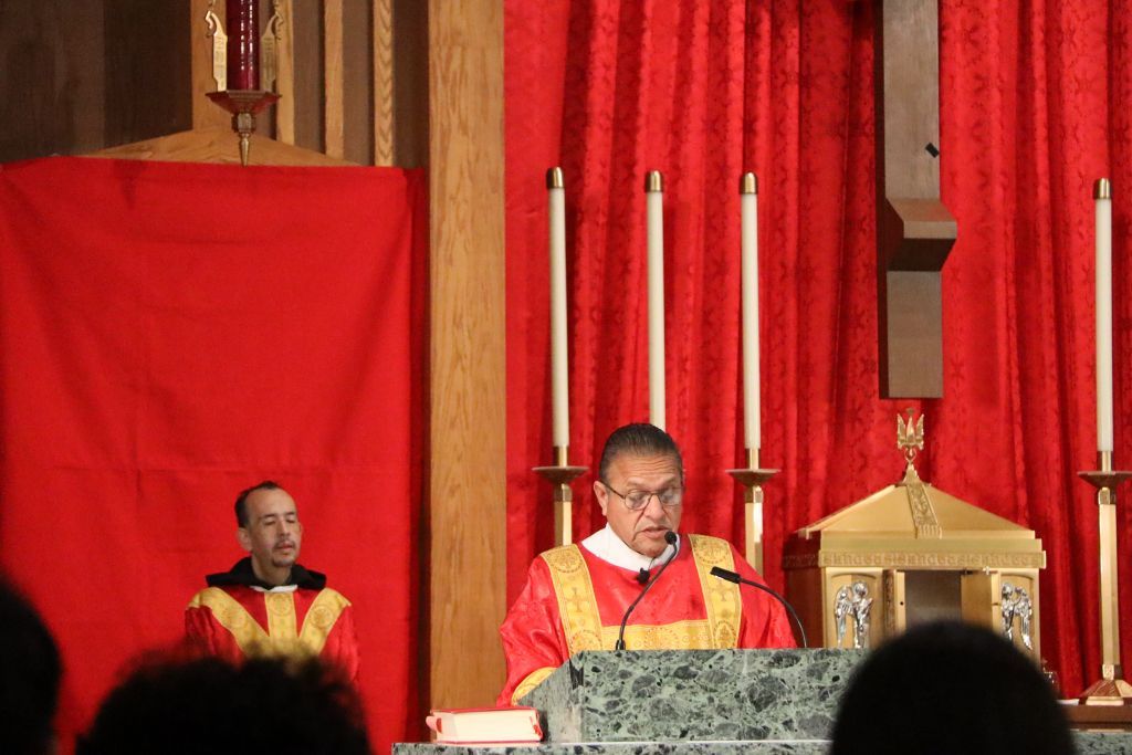Clergy in red and gold vestments at an altar with candles and a red draped backdrop