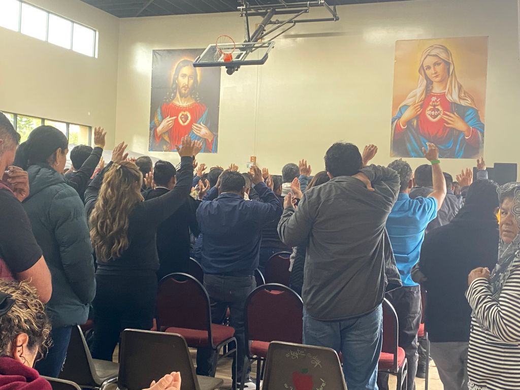 Crowd standing in a hall, raising hands toward two large religious portraits at the front.