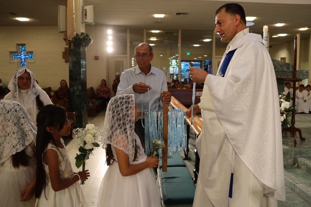 Children in white dresses receive communion from a priest in a church during a ceremony.