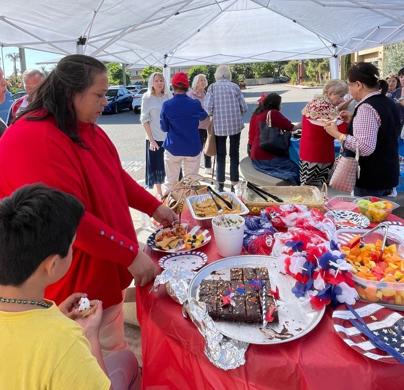 People browsing a red table at an outdoor fair under a white canopy, with patriotic decorations and treats