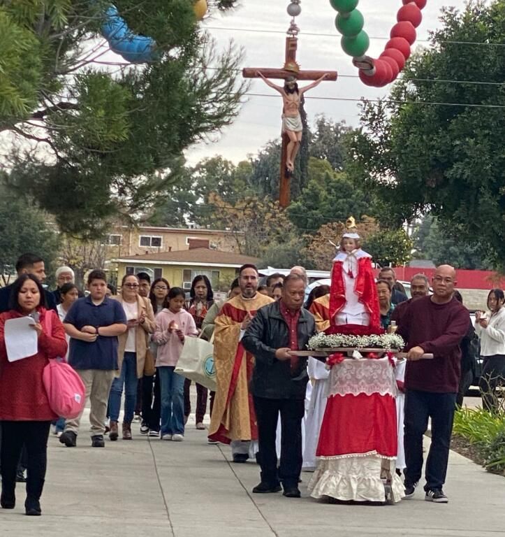 Religious procession outdoors with a crucifix overhead and people carrying a decorated statue along a sidewalk