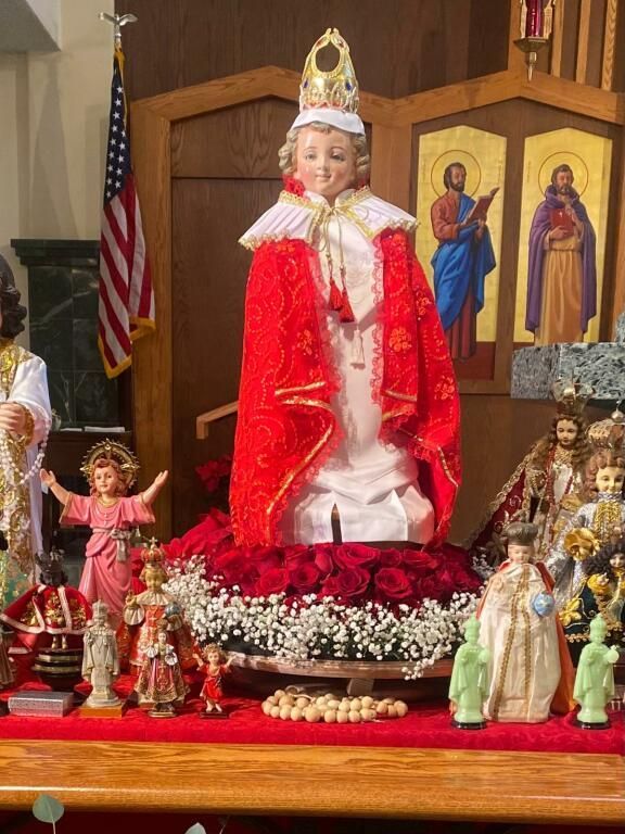 Catholic shrine display with a crowned saint statue in red robes, surrounded by smaller figures and flowers