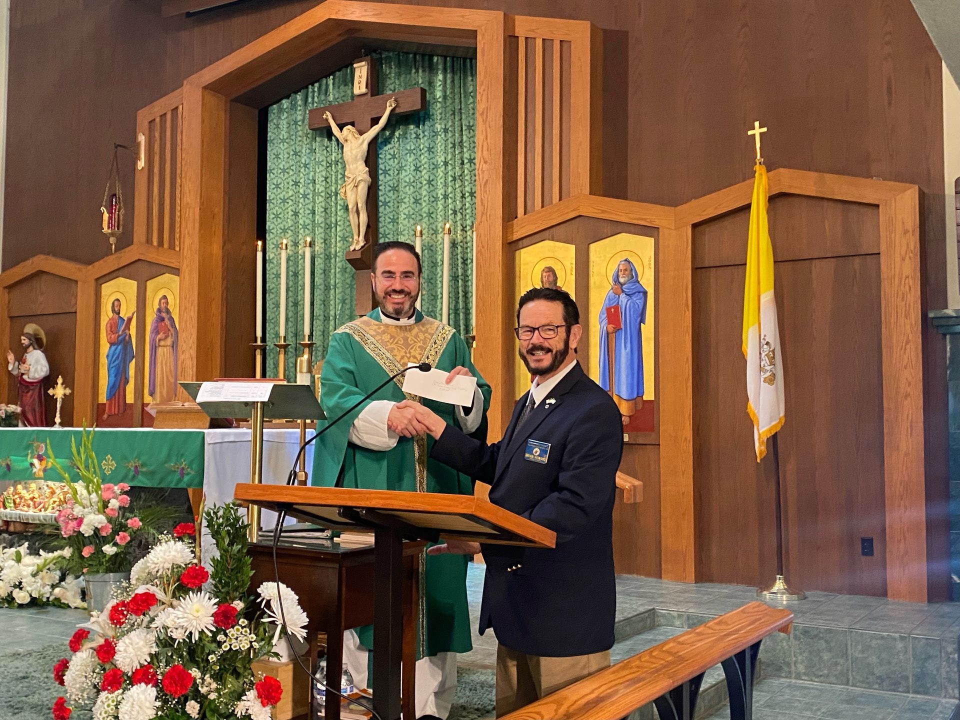 Two men at a church lectern, one in green vestments holding a certificate, with crucifix and flowers behind them