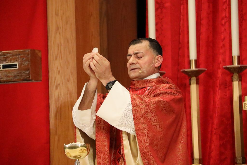 Priest in red vestments raises a chalice during a religious ceremony before red drapery and candles