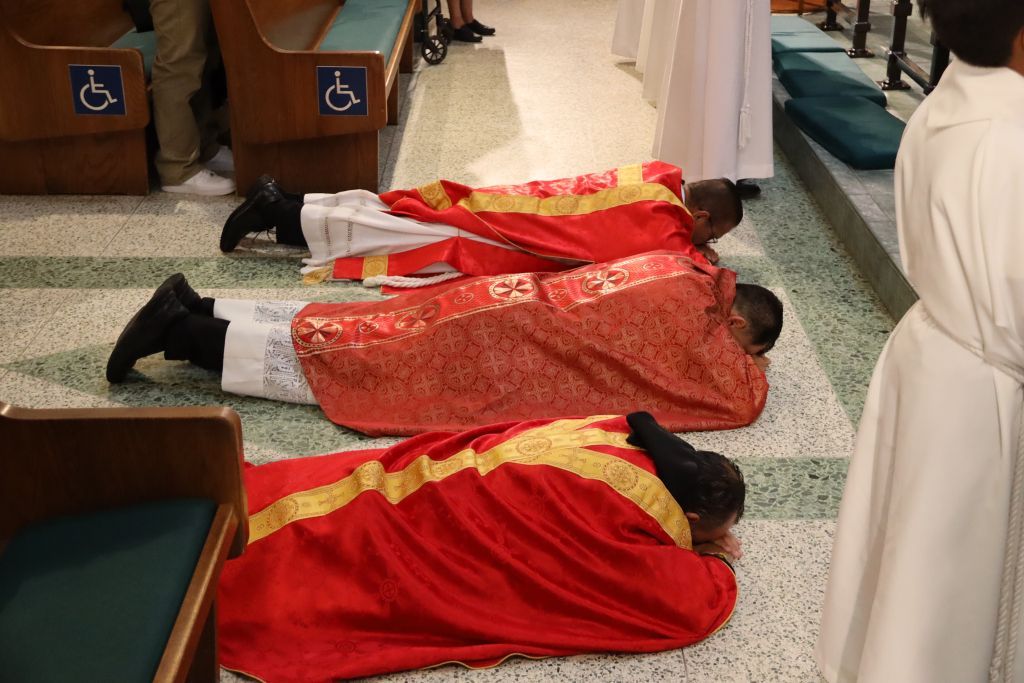 People lying face down on a floor, covered with red cloths in a room with prayer mats and chairs