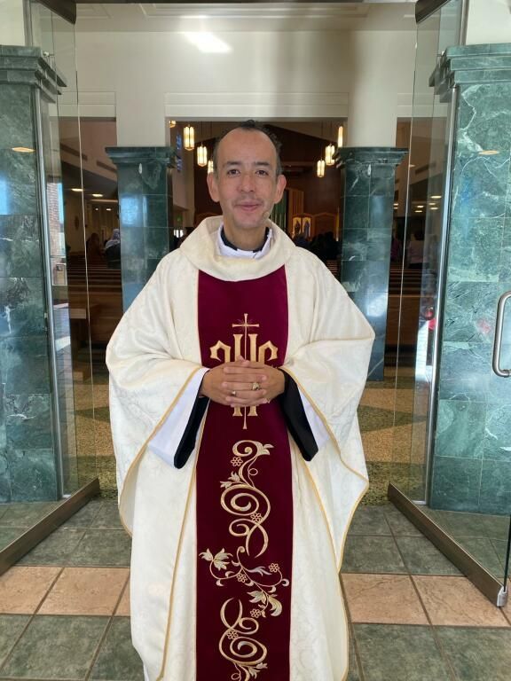 Clergyman in white vestments with maroon stole standing in a church entrance, hands clasped.