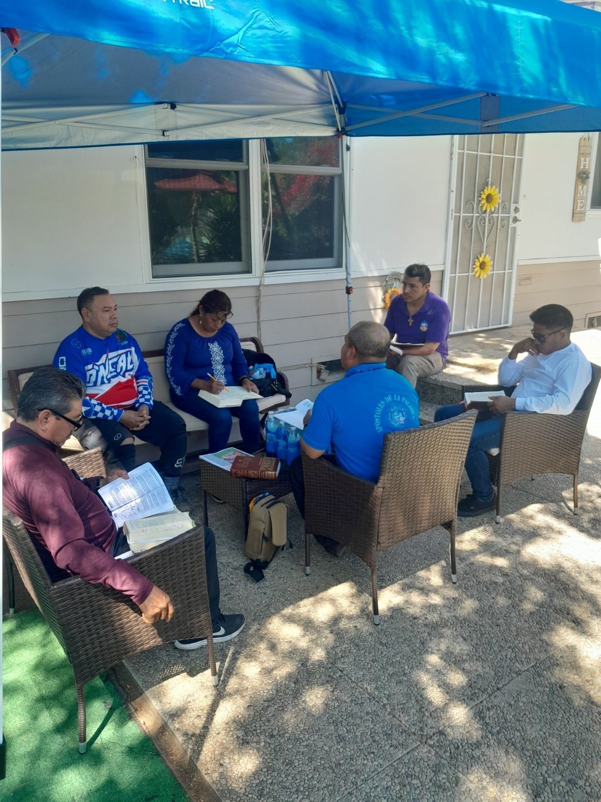 People seated in a shaded outdoor circle under a blue canopy, in discussion around a table.