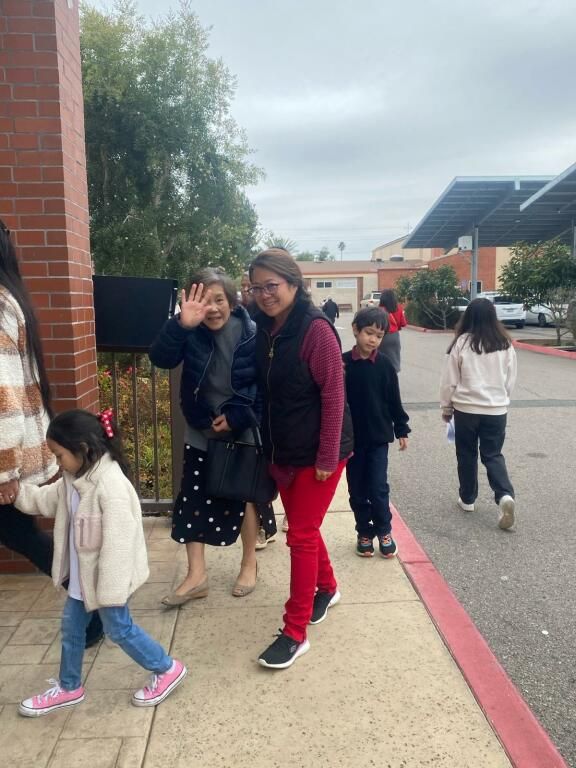 Children walking outside a school building, one waving at the camera, on a sidewalk by a curb