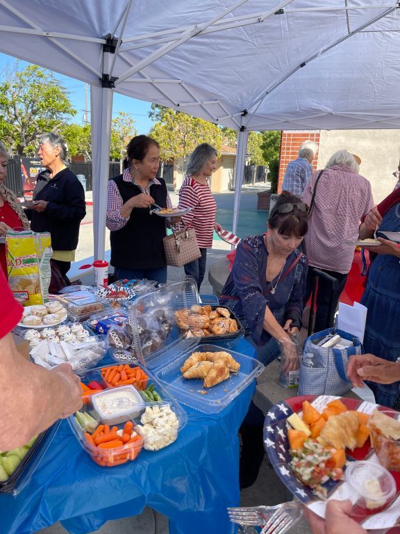 People serving and eating food at a blue-clothed outdoor buffet under a white canopy