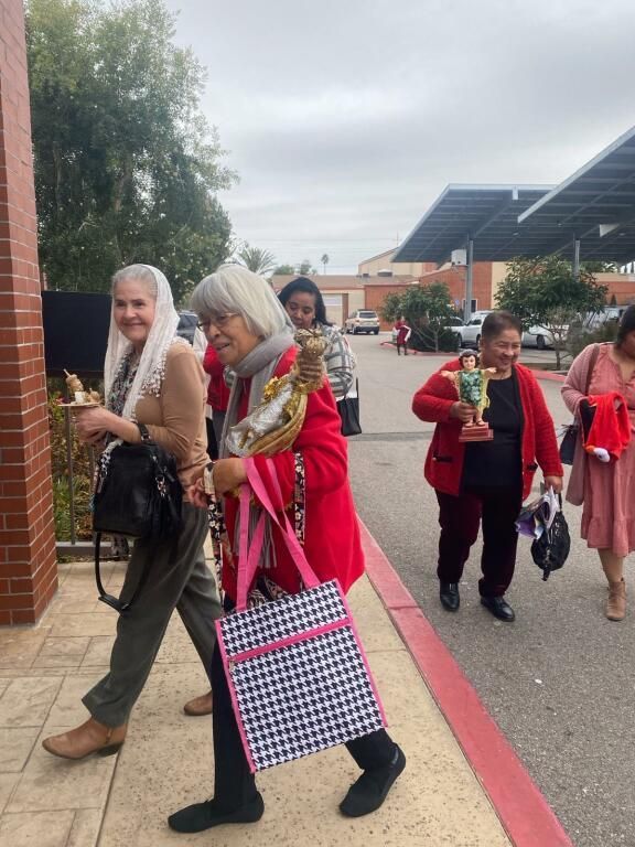 People walking outdoors near a brick building, carrying bags and gifts on a red-paved path.