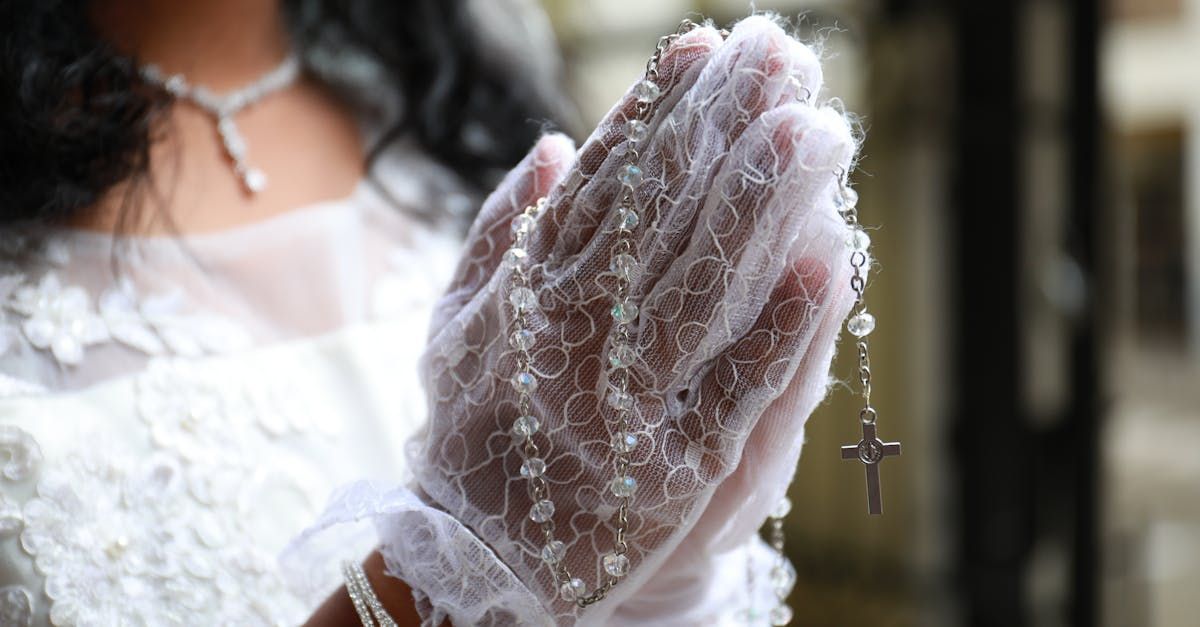 Lace-gloved hands clasped in prayer, with a white dress and rosary beads visible.